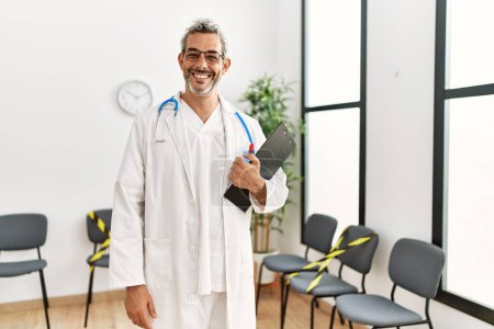 Téléchargez les photos : Moyen âge homme aux cheveux gris médecin souriant confiant tenant rapport médical à la salle d'attente clinique - en image libre de droit