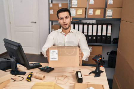 Photo for Young hispanic man with beard working at small business ecommerce holding box in shock face, looking skeptical and sarcastic, surprised with open mouth - Royalty Free Image