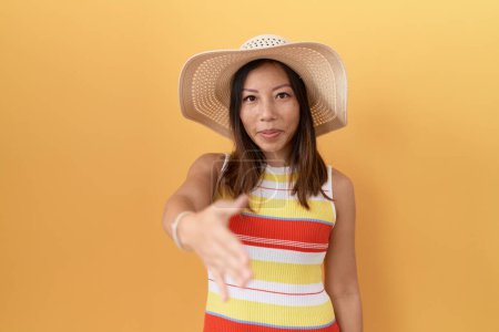 Foto de Mujer china de mediana edad con sombrero de verano sobre fondo amarillo sonriendo amistoso ofreciendo apretón de manos como saludo y bienvenida. negocios exitosos. - Imagen libre de derechos