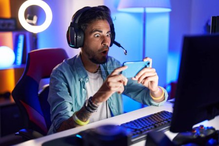 Photo for Young hispanic man with tattoos playing video games with smartphone scared and amazed with open mouth for surprise, disbelief face - Royalty Free Image