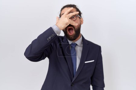 Foto de Hombre hispano con barba vistiendo traje y corbata asomándose en shock cubriendo cara y ojos con la mano, mirando a través de los dedos con expresión avergonzada. - Imagen libre de derechos