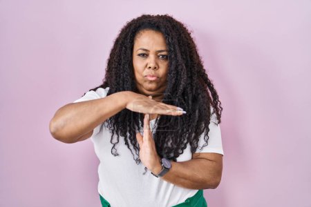 Foto de Más tamaño mujer hispana de pie sobre fondo rosa haciendo el gesto de tiempo de espera con las manos, rostro frustrado y grave - Imagen libre de derechos