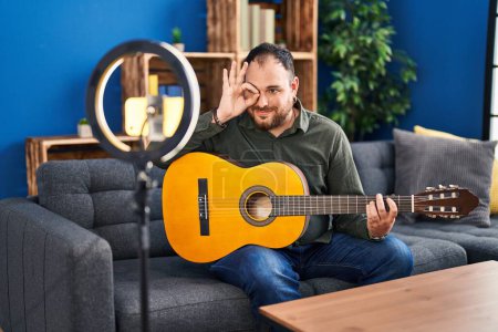 Photo for Plus size hispanic man with beard playing classic guitar at music studio recording himself smiling happy doing ok sign with hand on eye looking through fingers - Royalty Free Image