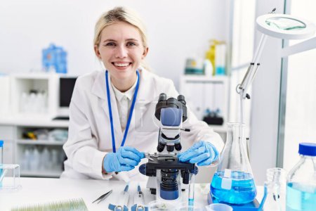 Photo for Young blonde woman scientist using microscope smiling at laboratory - Royalty Free Image