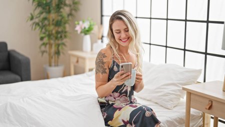Foto de Young blonde woman using smartphone drinking coffee at bedroom - Imagen libre de derechos