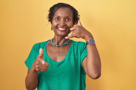 Téléchargez les photos : Femme africaine avec dreadlocks debout sur fond jaune souriant faire parler au téléphone geste et vous pointant du doigt. Appelle-moi. - en image libre de droit