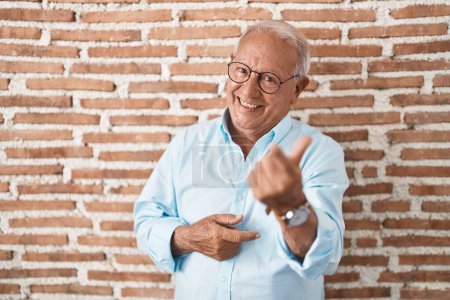 Foto de Hombre mayor con el pelo gris de pie sobre la pared de ladrillos señas vienen aquí gesto con la mano invitando a la bienvenida feliz y sonriente - Imagen libre de derechos