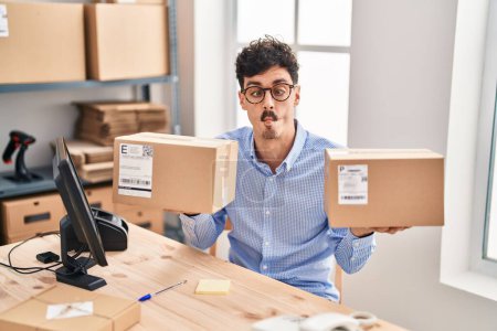 Photo for Hispanic man working at small business ecommerce holding packages making fish face with mouth and squinting eyes, crazy and comical. - Royalty Free Image