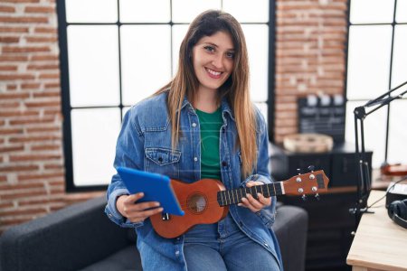 Photo for Young hispanic woman musician using touchpad holding ukulele at music studio - Royalty Free Image
