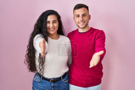 Foto de Joven pareja hispana de pie sobre fondo rosa sonriente amistoso ofreciendo apretón de manos como saludo y bienvenida. negocios exitosos. - Imagen libre de derechos