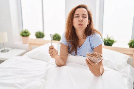 Téléchargez les photos : Femme brune prenant le petit déjeuner dans le lit gonflant les joues avec un visage drôle. bouche gonflée d'air, attrapant l'air. - en image libre de droit