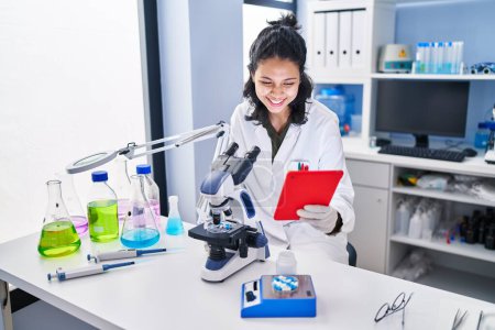 Photo for Young latin woman scientist using microscope and touchpad at laboratory - Royalty Free Image