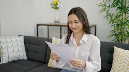 Photo for Young beautiful hispanic woman reading document sitting on sofa at home - Royalty Free Image