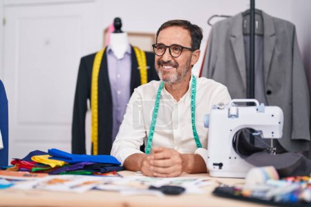 Foto de Middle age man tailor smiling confident sitting on table at tailor shop - Imagen libre de derechos