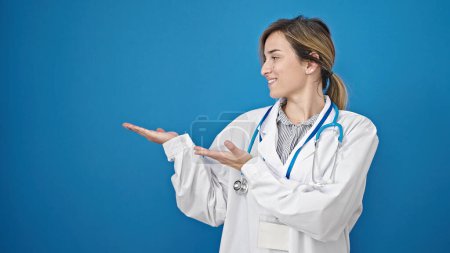 Photo for Young blonde woman doctor smiling confident presenting over isolated blue background - Royalty Free Image