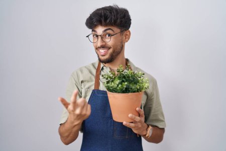 Téléchargez les photos : Homme arabe avec barbe tenant pot de plante verte signe de venir ici geste avec la main invitant accueillant heureux et souriant - en image libre de droit