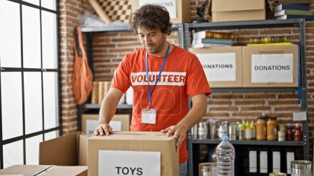 Téléchargez les photos : Jeune homme hispanique volontaire emballant des jouets sur le paquet de dons au centre de bienfaisance - en image libre de droit