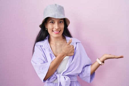 Foto de Mujer hispana joven de pie sobre fondo rosa con sombrero mostrando la palma de la mano y haciendo buen gesto con los pulgares hacia arriba, sonriendo feliz y alegre - Imagen libre de derechos