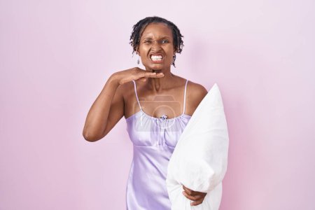 Photo for African woman with dreadlocks wearing pajama hugging pillow cutting throat with hand as knife, threaten aggression with furious violence - Royalty Free Image