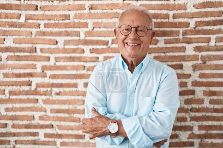 Foto de Hombre mayor con el pelo gris de pie sobre la pared de ladrillos cara feliz sonriendo con los brazos cruzados mirando a la cámara. persona positiva. - Imagen libre de derechos