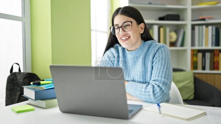 Téléchargez les photos : Jeune étudiante hispanique utilisant un ordinateur portable souriant à l'université de la bibliothèque - en image libre de droit