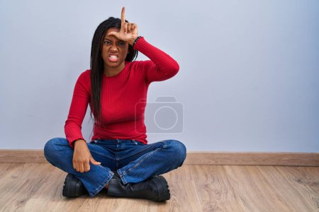 Photo for Young african american with braids sitting on the floor at home making fun of people with fingers on forehead doing loser gesture mocking and insulting. - Royalty Free Image