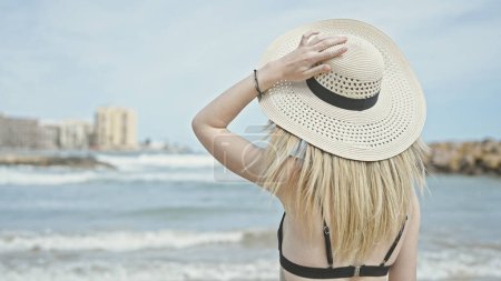 Téléchargez les photos : Jeune femme blonde touriste portant un bikini et un chapeau d'été debout vers l'arrière à la plage - en image libre de droit