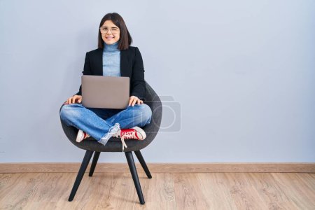 Téléchargez les photos : Jeune femme hispanique assis sur la chaise en utilisant ordinateur portable clin d'oeil en regardant la caméra avec une expression sexy, gai et heureux visage. - en image libre de droit