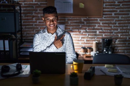 Photo for Young hispanic man working at the office at night cheerful with a smile of face pointing with hand and finger up to the side with happy and natural expression on face - Royalty Free Image