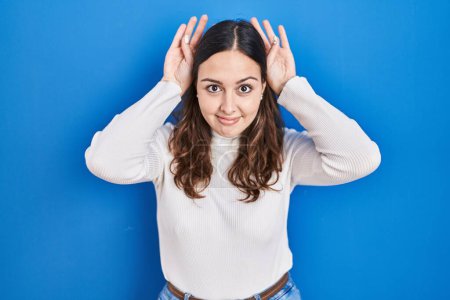Foto de Young hispanic woman standing over blue background doing bunny ears gesture with hands palms looking cynical and skeptical. easter rabbit concept. - Imagen libre de derechos