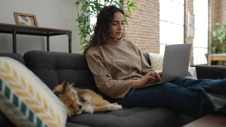 Foto de Mujer hispana joven con perro usando portátil sentado en el sofá en casa - Imagen libre de derechos