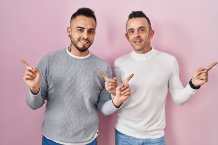 Téléchargez les photos : Couple homosexuel debout sur fond rose souriant confiant pointant avec les doigts vers différentes directions. espace de copie pour la publicité - en image libre de droit