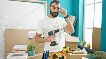 Téléchargez les photos : Jeune homme hispanique réparateur tenant la perceuse portant des lunettes de sécurité à la nouvelle maison - en image libre de droit