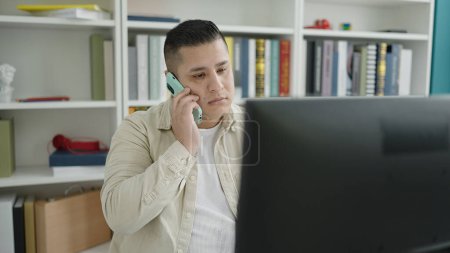 Téléchargez les photos : Young hispanic man student using computer talking on smartphone at library university - en image libre de droit