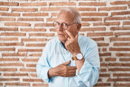 Photo for Senior man with grey hair standing over bricks wall pointing to the eye watching you gesture, suspicious expression - Royalty Free Image