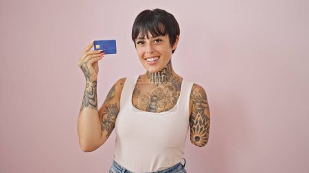 Spanierin mit amputiertem Arm lächelt selbstbewusst und hält Kreditkarte vor isoliertem rosa Hintergrund