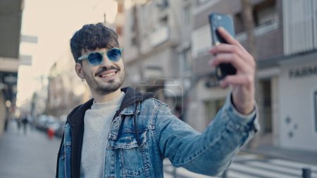 Foto de Joven hombre hispano sonriendo confiado haciendo selfie con smartphone en la calle - Imagen libre de derechos
