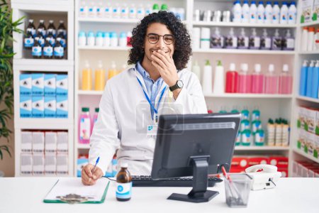 Téléchargez les photos : Homme hispanique aux cheveux bouclés travaillant à la pharmacie drugstore rire et rire embarrassé couvrant la bouche avec les mains, commérages et concept de scandale - en image libre de droit