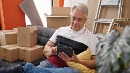 Téléchargez les photos : Homme d'âge moyen avec les cheveux gris assis sur le canapé en utilisant la tablette à la nouvelle maison - en image libre de droit