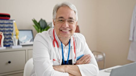Téléchargez les photos : Homme d'âge moyen avec cheveux gris médecin souriant confiant avec les bras croisés à la clinique - en image libre de droit