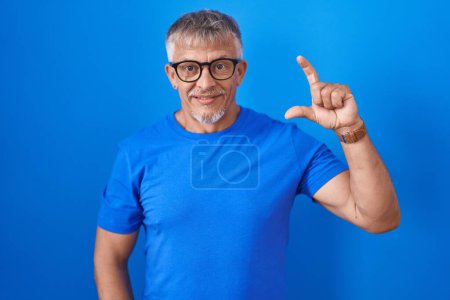 Téléchargez les photos : Homme hispanique avec les cheveux gris debout sur fond bleu geste souriant et confiant avec la main faisant signe de petite taille avec les doigts regardant et la caméra. concept de mesure. - en image libre de droit