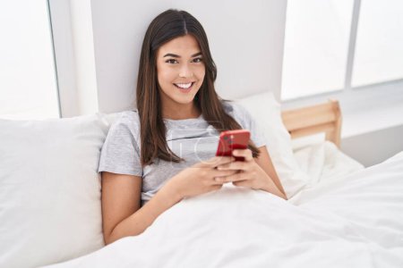 Téléchargez les photos : Jeune belle femme hispanique utilisant smartphone assis sur le lit dans la chambre - en image libre de droit