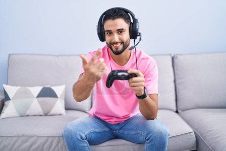 Téléchargez les photos : Jeune homme hispanique jouant contrôleur de tenue de jeu vidéo assis sur le canapé signe viennent ici geste avec la main invitant accueillant heureux et souriant - en image libre de droit