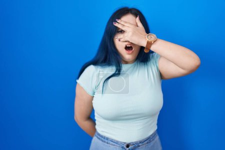 Photo for Young modern girl with blue hair standing over blue background peeking in shock covering face and eyes with hand, looking through fingers with embarrassed expression. - Royalty Free Image