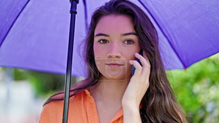 Foto de Joven mujer hispana hermosa sosteniendo paraguas hablando en el teléfono inteligente en el parque - Imagen libre de derechos