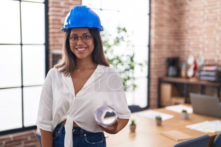 Photo for Hispanic young woman wearing architect hardhat at office looking positive and happy standing and smiling with a confident smile showing teeth - Royalty Free Image