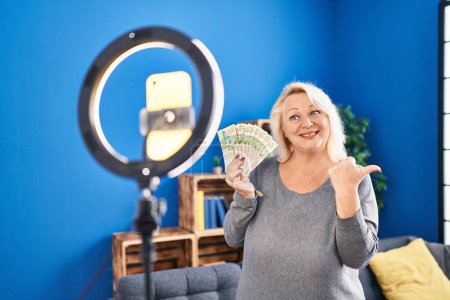 Photo for Middle age caucasian woman recording vlog tutorial with smartphone at home holding hong kong dollars pointing thumb up to the side smiling happy with open mouth - Royalty Free Image