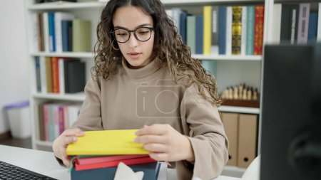 Téléchargez les photos : Jeune belle étudiante hispanique à la recherche de livres à la bibliothèque de l'université - en image libre de droit
