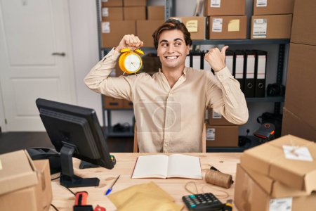 Photo for Young man working at small business ecommerce holding alarm clock pointing thumb up to the side smiling happy with open mouth - Royalty Free Image