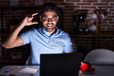Photo for Hispanic man with beard using laptop at night smiling and confident gesturing with hand doing small size sign with fingers looking and the camera. measure concept. - Royalty Free Image
